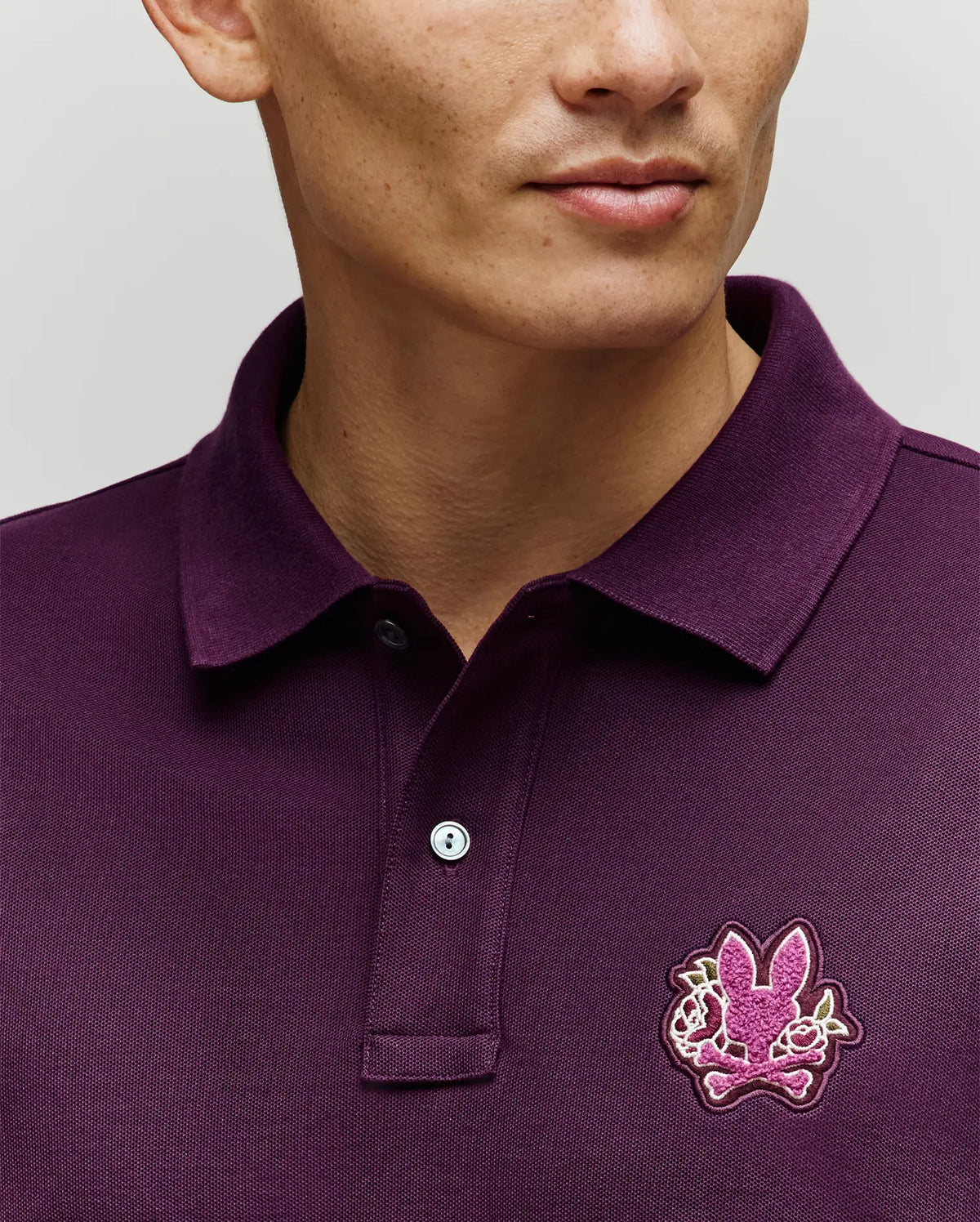 A person is shown wearing the Psycho Bunny MENS AUGUSTE PIQUE POLO SHIRT (MT0100051) in dark purple, featuring a pink chenille bunny patch on the chest. The image highlights the upper torso and part of the face against a neutral background.