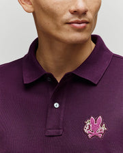 A person is shown wearing the Psycho Bunny MENS AUGUSTE PIQUE POLO SHIRT (MT0100051) in dark purple, featuring a pink chenille bunny patch on the chest. The image highlights the upper torso and part of the face against a neutral background.