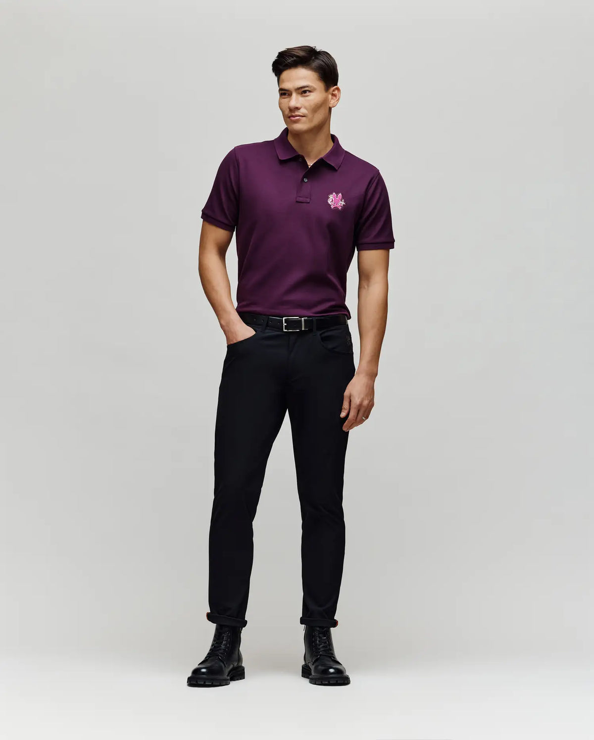 A man stands against a light background wearing the Psycho Bunny MENS AUGUSTE PIQUE POLO SHIRT - MT0100051 in dark purple with a chenille bunny patch on the left chest, black pants, belt, and boots. He has his hands in his pockets and is slightly smiling.