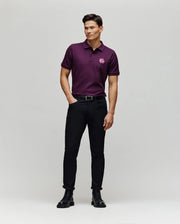 A man stands against a light background wearing the Psycho Bunny MENS AUGUSTE PIQUE POLO SHIRT - MT0100051 in dark purple with a chenille bunny patch on the left chest, black pants, belt, and boots. He has his hands in his pockets and is slightly smiling.