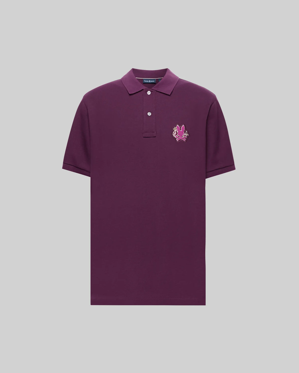 The Psycho Bunny MENS AUGUSTE PIQUE POLO SHIRT (MT0100051) is a short-sleeved, dark purple pima cotton polo with a collar, two white buttons, and a pink-and-white embroidered crest on the left chest, shown on a light gray background.