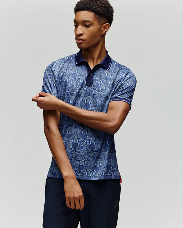 A young man wearing the Psycho Bunny MENS REEVES SPORT POLO SHIRT (MT0100053) and dark pants stands against a plain light background, looking to the side as he adjusts his left sleeve with his right hand.