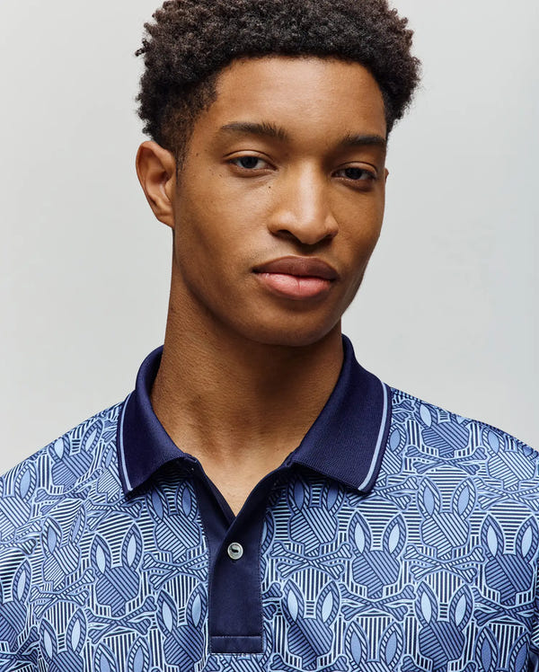 A young man with short curly hair wears the Psycho Bunny MENS REEVES SPORT POLO SHIRT (MT0100053) featuring a breathable Bunny print and dark collar, standing against a plain light background, looking slightly toward the camera with a neutral expression.