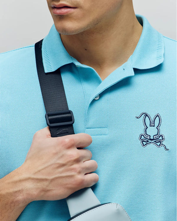 A man wears the Psycho Bunny MENS CYRUS PIQUE POLO - MT0100056 in light blue, made from premium Pima cotton with a rabbit logo on the chest, holding a black strap of a gray bag; only his lower face and upper body are visible.