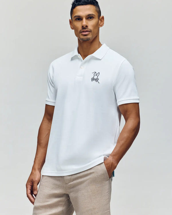A man wears the Psycho Bunny MENS CYRUS PIQUE POLO - MT0100056, a white short-sleeve 100% Pima cotton polo with a rabbit logo on the chest, paired with light beige pants, against a plain light background.