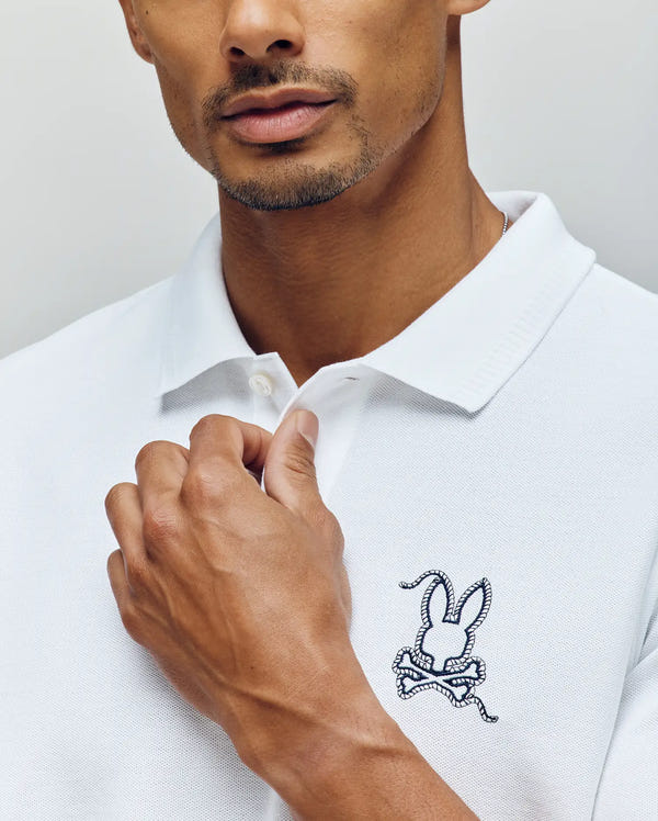 A man wearing the Psycho Bunny MENS CYRUS PIQUE POLO - MT0100056 in a bold color, made from 100% Pima cotton, featuring a large embroidered bunny head and crossed bones on the chest, holding the shirt collar with one hand.