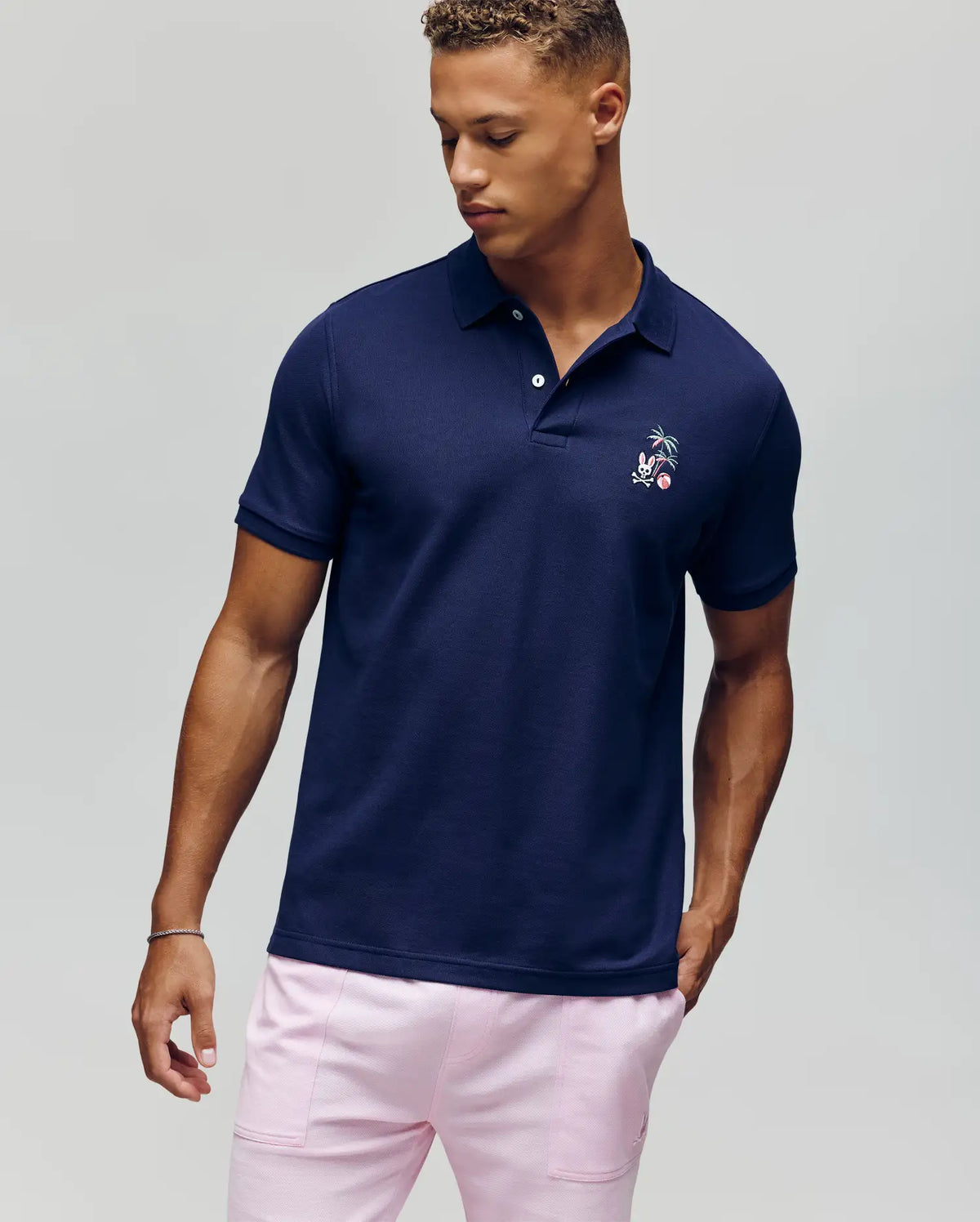 A man stands against a plain light background, looking down to his left, wearing the Psycho Bunny MENS SONNY PIQUE POLO - MT0100094 in navy blue with subtle chest embroidery and light pink pants.