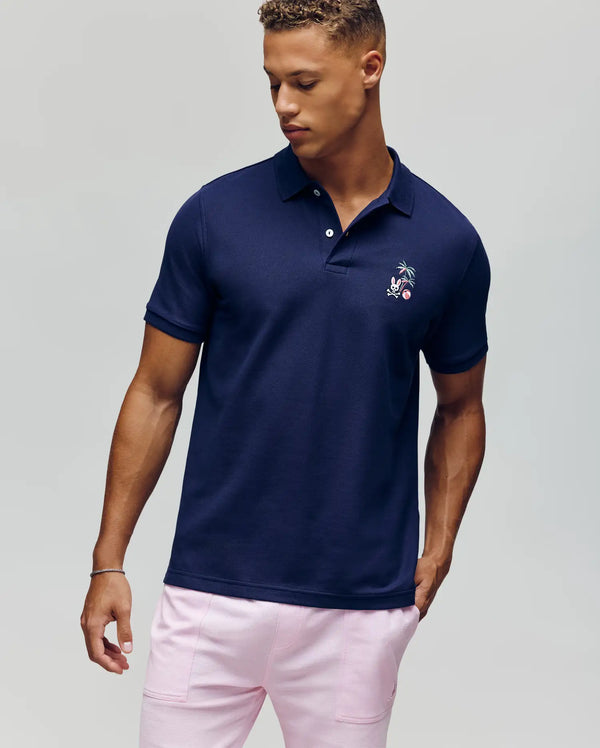 A man stands against a plain light background, looking down to his left, wearing the Psycho Bunny MENS SONNY PIQUE POLO - MT0100094 in navy blue with subtle chest embroidery and light pink pants.