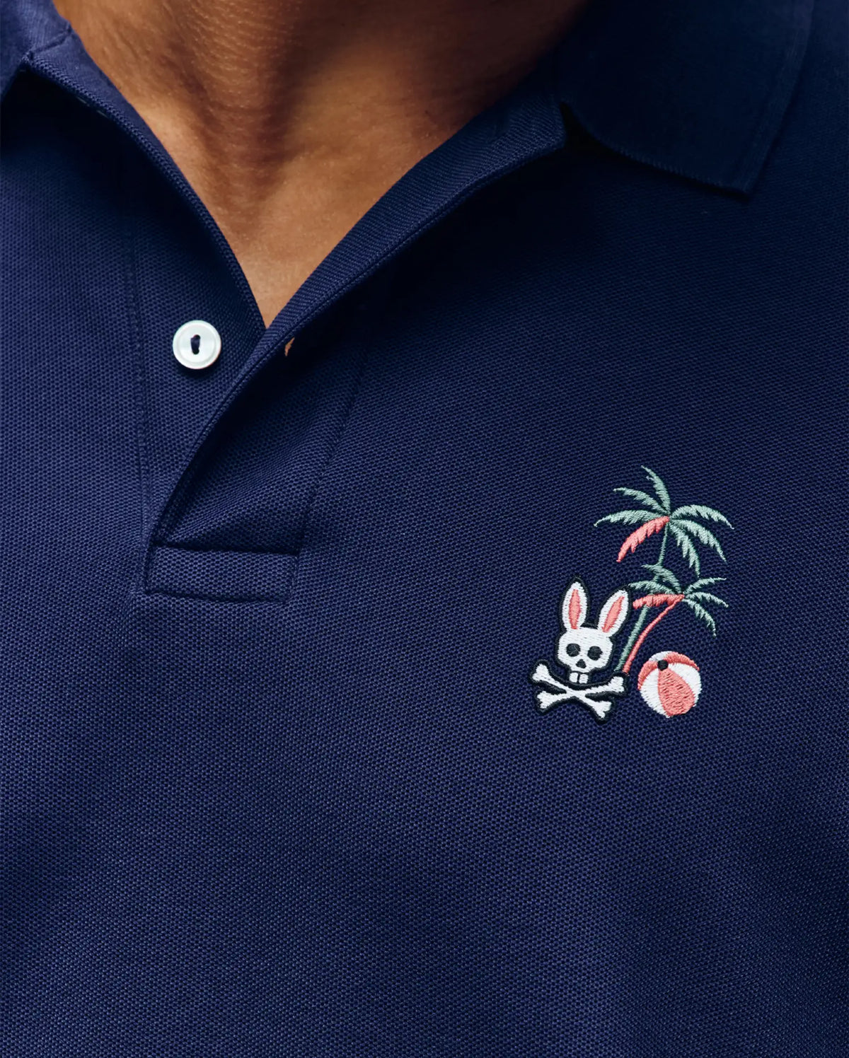 Close-up of the Psycho Bunny MENS SONNY PIQUE POLO (MT0100094) in navy Pima cotton, featuring a white button and an embroidered rabbit skull and crossbones, palm tree, and beach ball on the left chest.