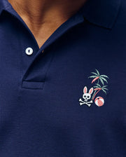 Close-up of the Psycho Bunny MENS SONNY PIQUE POLO (MT0100094) in navy Pima cotton, featuring a white button and an embroidered rabbit skull and crossbones, palm tree, and beach ball on the left chest.