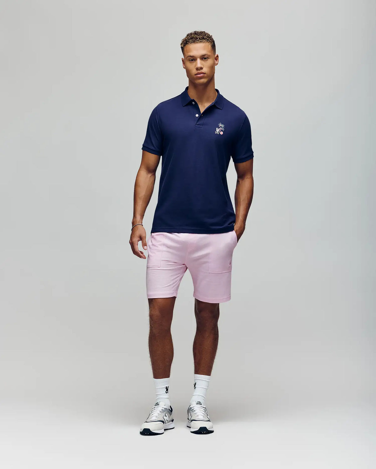 A man with short curly hair and a neutral look wears the Psycho Bunny MENS SONNY PIQUE POLO - MT0100094 in navy, paired with light pink shorts, white crew socks, and white sneakers against a plain background.