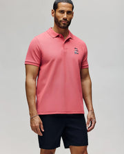 A man with short hair and a beard stands against a plain background wearing the Psycho Bunny MENS SONNY PIQUE POLO - MT0100094 in pink, featuring subtle chest embroidery, paired with black shorts and a neutral expression.