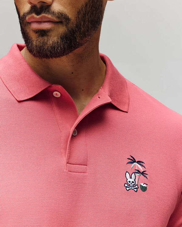 A man wears the Psycho Bunny MENS SONNY PIQUE POLO (MT0100094), a pink Pima cotton shirt with a classic collar, two buttons, and chest embroidery featuring a bunny, crossed bones, palm trees, and a green ball.