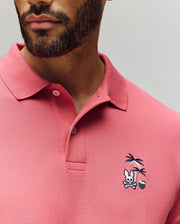 A man wears the Psycho Bunny MENS SONNY PIQUE POLO (MT0100094), a pink Pima cotton shirt with a classic collar, two buttons, and chest embroidery featuring a bunny, crossed bones, palm trees, and a green ball.