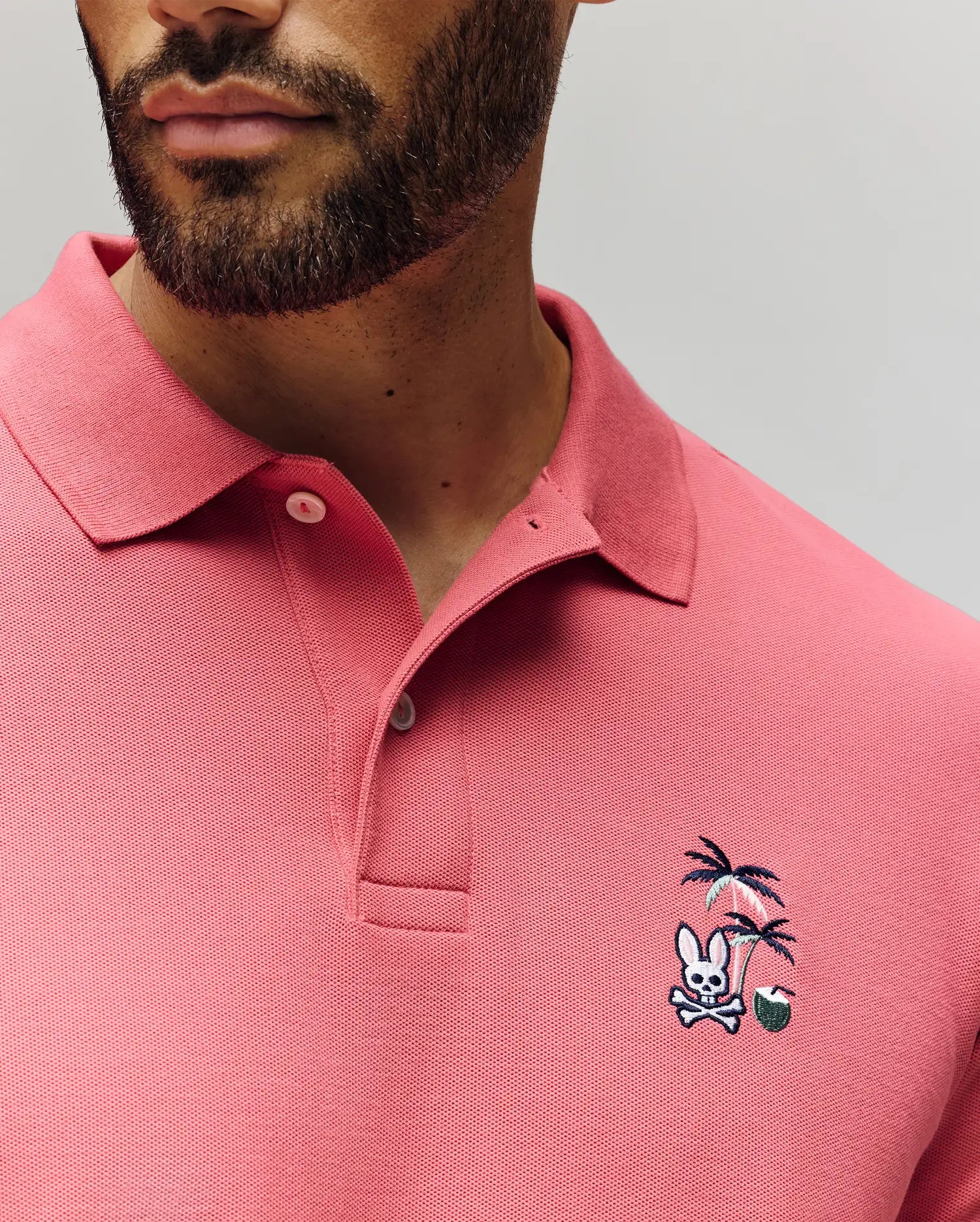 A man wears the Psycho Bunny MENS SONNY PIQUE POLO (MT0100094), a pink Pima cotton shirt with a classic collar, two buttons, and chest embroidery featuring a bunny, crossed bones, palm trees, and a green ball.