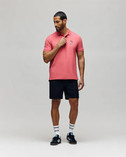 A man stands against a plain background wearing the Psycho Bunny MENS SONNY PIQUE POLO - MT0100094 in pink, black shorts, striped white socks, and black Adidas sneakers, looking down and touching his collar with one hand.