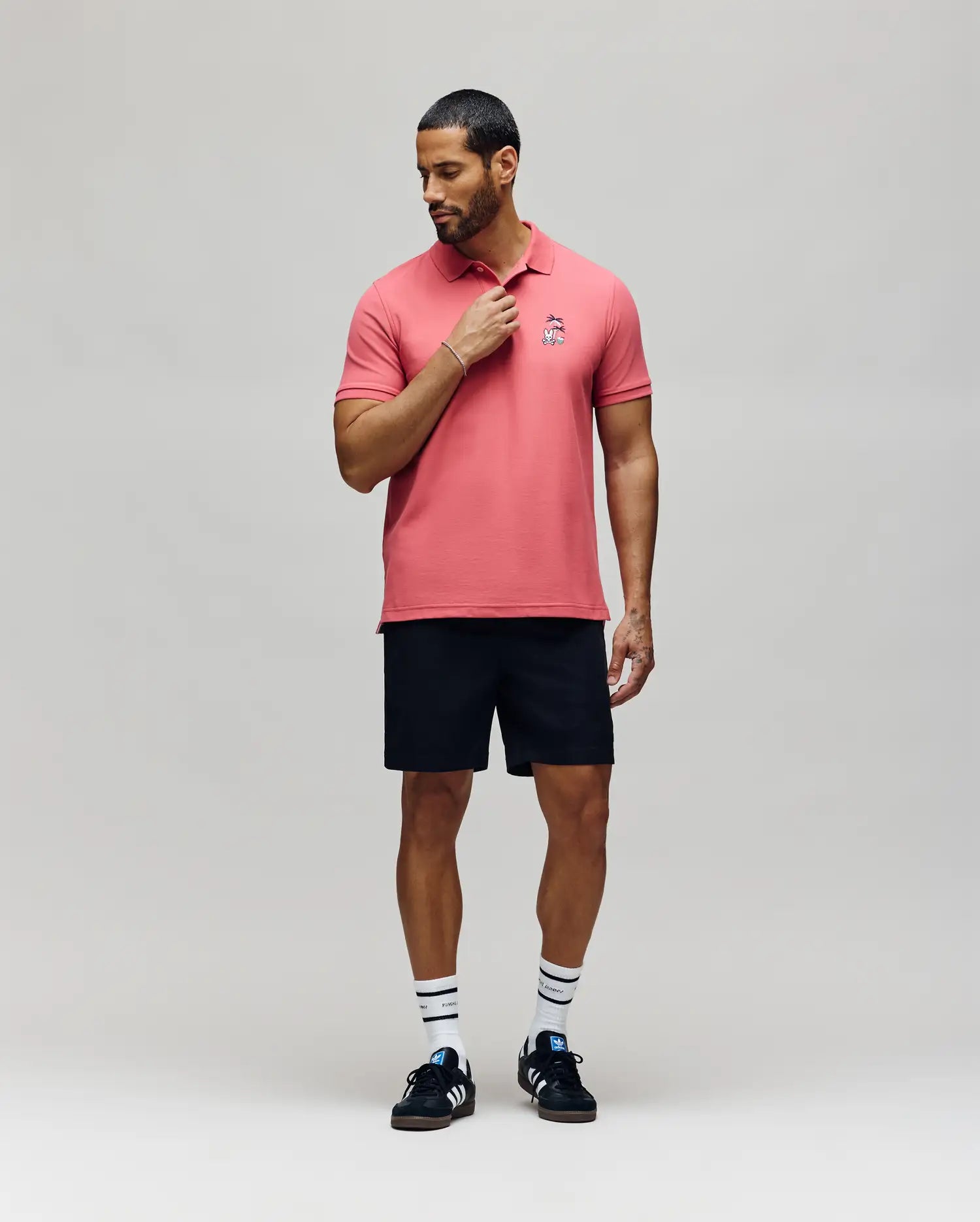A man stands against a plain background wearing the Psycho Bunny MENS SONNY PIQUE POLO - MT0100094 in pink, black shorts, striped white socks, and black Adidas sneakers, looking down and touching his collar with one hand.