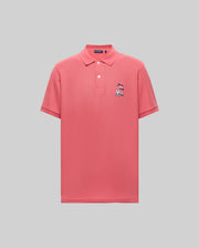 The Psycho Bunny MENS SONNY PIQUE POLO (MT0100094) is a coral pink Pima cotton short-sleeve polo with a collar, three buttons, and a small cartoon chest embroidery on the left, shown on a light gray background.