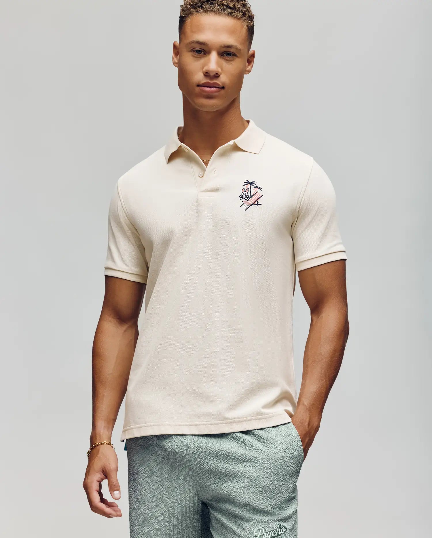 A young man with short curly hair wears a Psycho Bunny MENS SONNY PIQUE POLO - MT0100094 in cream, featuring subtle chest embroidery, paired with light green textured shorts against a plain light gray background.