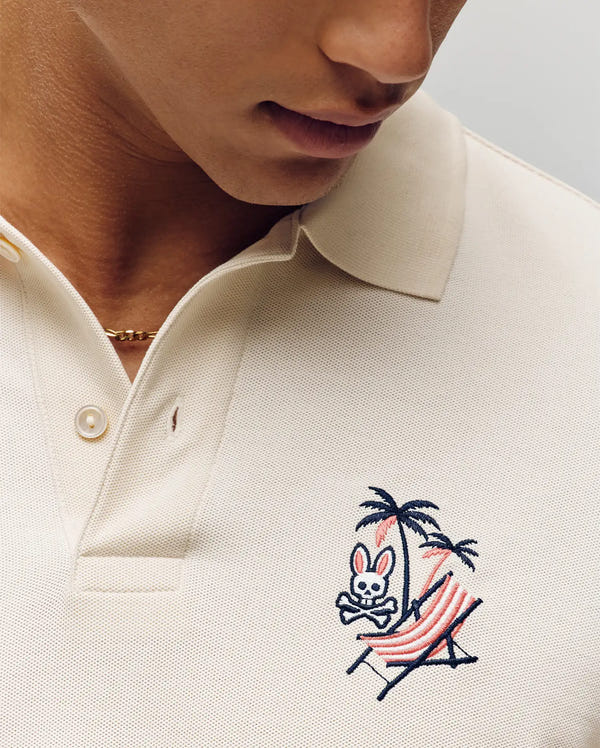 A person relaxes on a striped hammock under a palm tree, wearing the Psycho Bunny MENS SONNY PIQUE POLO (MT0100094), cream-colored Pima cotton with signature bunny skull embroidery, finished with a thin gold chain necklace.