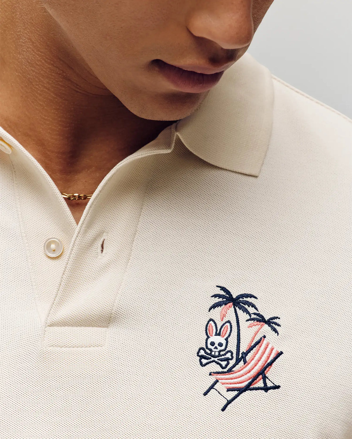 A person relaxes on a striped hammock under a palm tree, wearing the Psycho Bunny MENS SONNY PIQUE POLO (MT0100094), cream-colored Pima cotton with signature bunny skull embroidery, finished with a thin gold chain necklace.