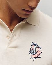 A person relaxes on a striped hammock under a palm tree, wearing the Psycho Bunny MENS SONNY PIQUE POLO (MT0100094), cream-colored Pima cotton with signature bunny skull embroidery, finished with a thin gold chain necklace.