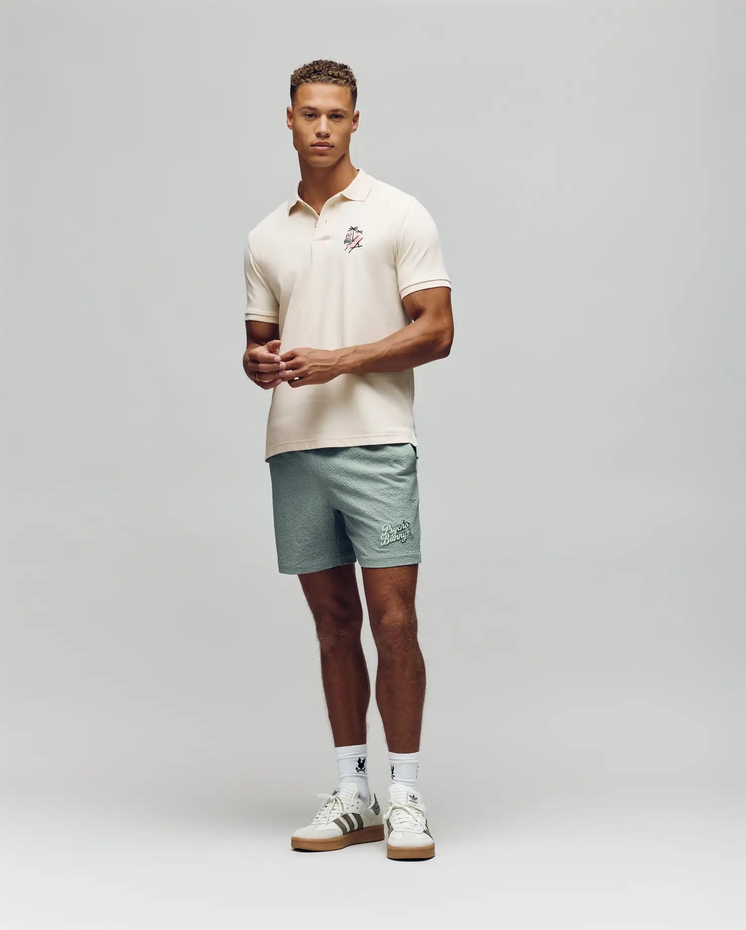 A man faces forward wearing the Psycho Bunny MENS SONNY PIQUE POLO (MT0100094) in cream, paired with light green shorts, white socks, and white sneakers with brown soles against a plain light grey background.