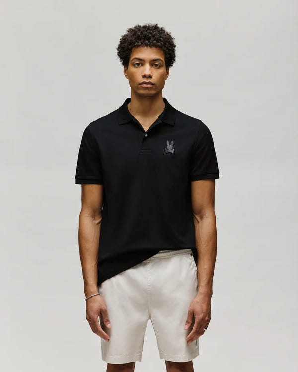 A man with curly hair wears the Psycho Bunny MENS PATRICK POLO - MT0100095 in black and light-colored shorts, standing against a plain, light grey background.