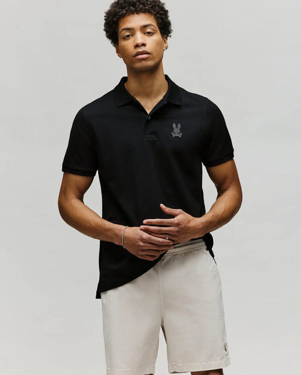 A man wearing the Psycho Bunny MENS PATRICK POLO (MT0100095), a black 100% cotton polo with a small chest emblem, and light shorts stands against a plain light gray background, looking forward with a neutral expression and one hand on his stomach.