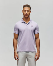 A young man faces forward wearing the Psycho Bunny MENS PATRICK POLO - MT0100095 in light purple with a bunny logo on the left chest and light gray pants, set against a plain light background.