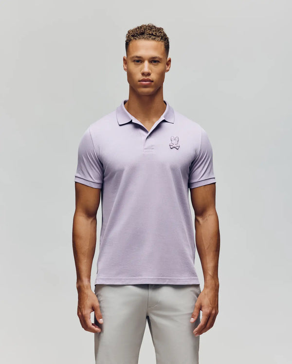 A young man faces forward wearing the Psycho Bunny MENS PATRICK POLO - MT0100095 in light purple with a bunny logo on the left chest and light gray pants, set against a plain light background.