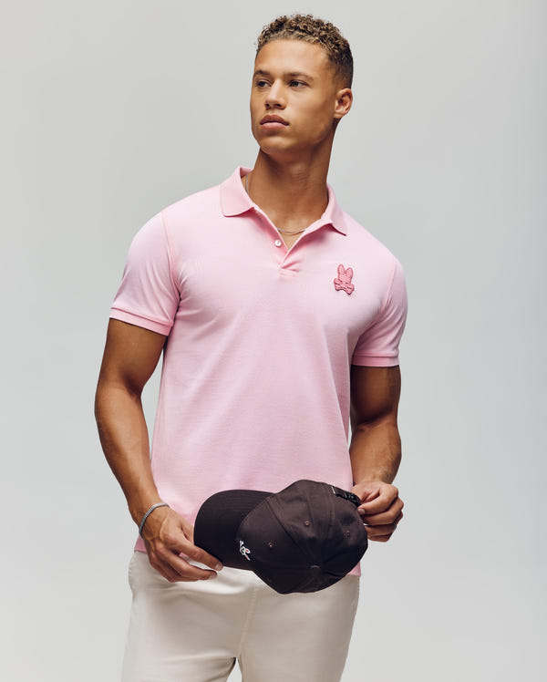 A young man with short curly hair and a fit build wears the Psycho Bunny MENS PATRICK POLO (MT0100095) in light pink, made from 100% cotton with the iconic bunny logo, standing against a plain background holding a black baseball cap.