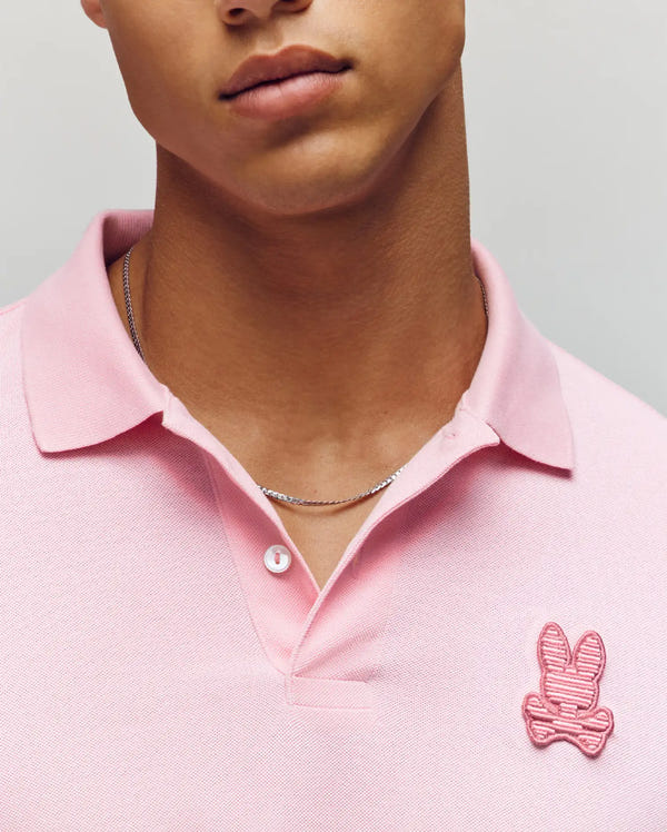 A person is shown from shoulders to lips wearing the Psycho Bunny MENS PATRICK POLO (MT0100095), a light pink 100% cotton knit shirt made in Peru with a pink bunny and crossbones logo on the chest, plus a thin silver chain necklace.