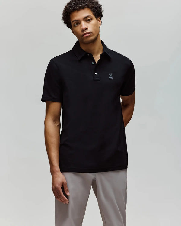 A man with short curly hair wears the Psycho Bunny MENS PHOENIX FASHION POLO - MT0100096 in black and light gray pants, standing against a plain light background with a neutral expression.