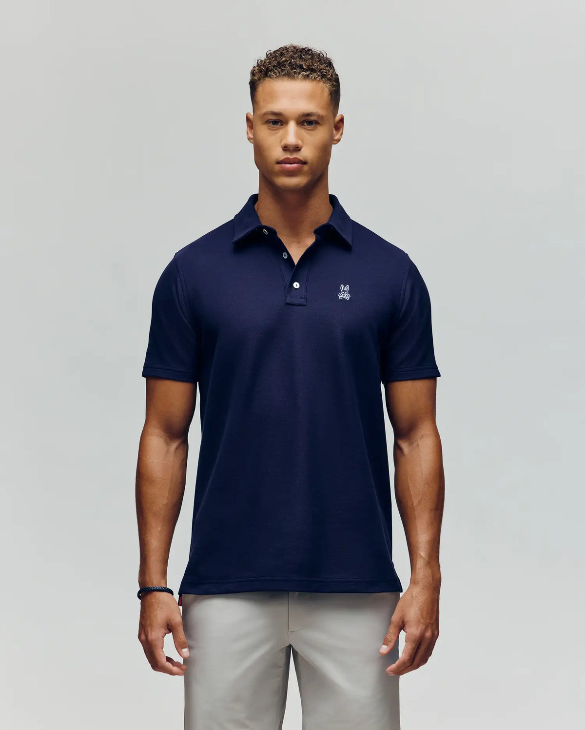 A man stands facing forward, wearing the Psycho Bunny MENS PHOENIX FASHION POLO - MT0100096 in navy blue with a small white logo on the chest and light-colored pants, against a plain light gray background.