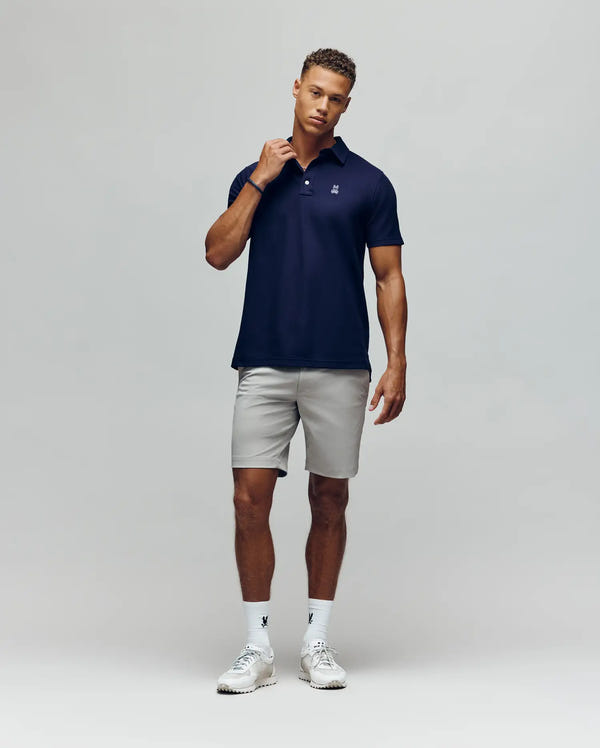 A man stands against a plain background wearing the Psycho Bunny MENS PHOENIX FASHION POLO - MT0100096 in navy, paired with light gray shorts, white socks, and white sneakers. He touches his collar and looks relaxed.