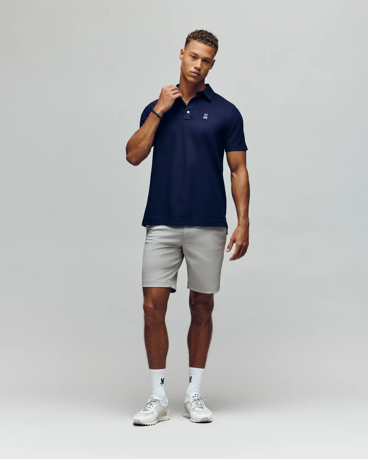 A man stands against a plain background wearing the Psycho Bunny MENS PHOENIX FASHION POLO - MT0100096 in navy, paired with light gray shorts, white socks, and white sneakers. He touches his collar and looks relaxed.