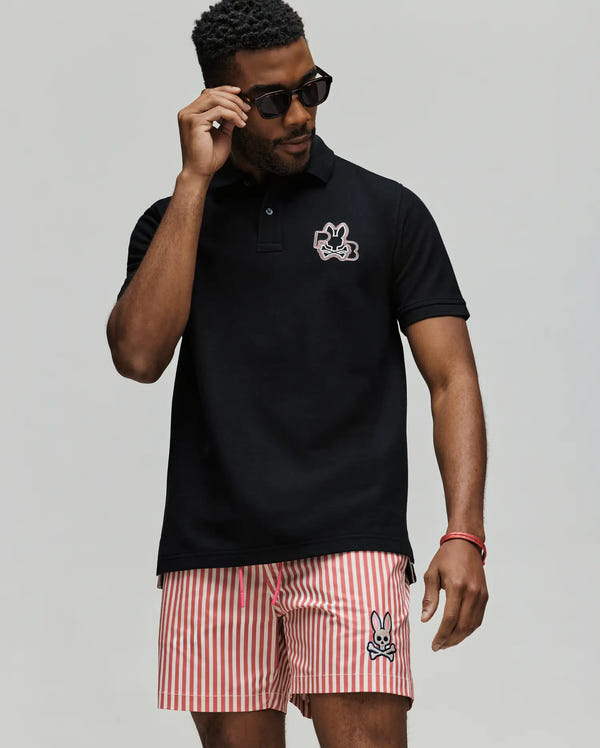 A man wears the Psycho Bunny MENS CARLO FASHION POLO - MT0100097, featuring a small bunny logo, paired with red-and-white striped shorts with matching logo, and sunglasses, standing against a plain light background.