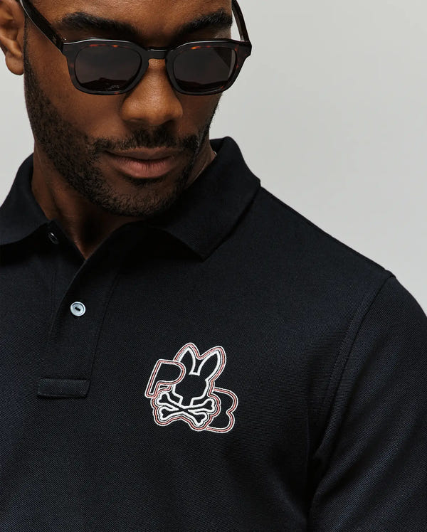 A man wearing dark sunglasses and a Psycho Bunny MENS CARLO FASHION POLO (MT0100097) with the bunny and crossbones logo on the chest is shown from the shoulders up against a plain background.