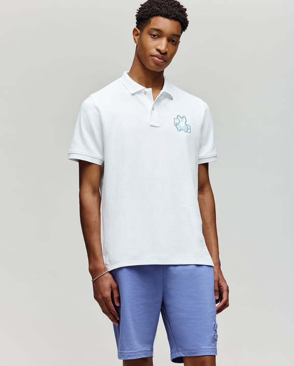 A young man stands against a plain background, wearing the Psycho Bunny MENS CARLO FASHION POLO - MT0100097, a white short-sleeved polo with a blue bunny logo, paired with light purple shorts. He looks relaxed with one hand in his pocket.