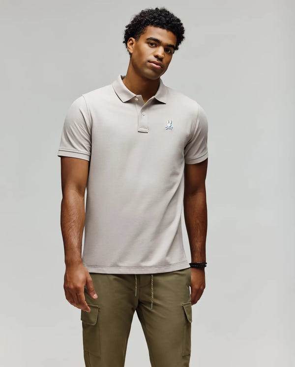 A young man with short curly hair wears the Psycho Bunny MENS CASSIAN PIQUE POLO - MT0100098 in light beige and olive cargo pants, standing against a plain light gray background, looking slightly to the side with a neutral expression.