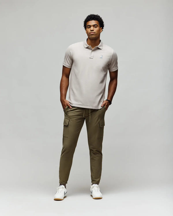 A man stands against a plain background wearing a light grey Psycho Bunny MENS CASSIAN PIQUE POLO - MT0100098, olive green cargo jogger pants, and white sneakers. His hands are in his pockets and he looks straight ahead with a neutral expression.