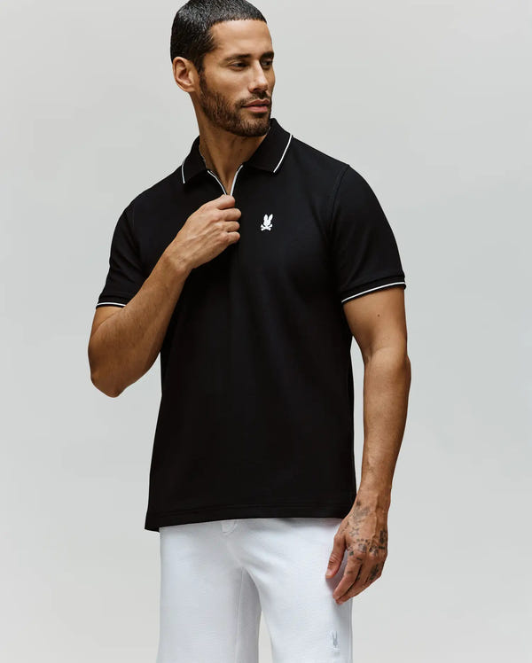 A man with short dark hair and a trimmed beard wears the Psycho Bunny MENS DANE ZIP PIQUE POLO - MT0100103 in black, featuring white trim and an embroidered Bunny logo, paired with white pants against a plain light background.