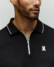 A man with a trimmed beard wears the Psycho Bunny MENS DANE ZIP PIQUE POLO (MT0100103), a black Pima cotton collared shirt featuring a white zipper and an embroidered Bunny logo. The sporty image is cropped from the shoulders to just above the mouth.