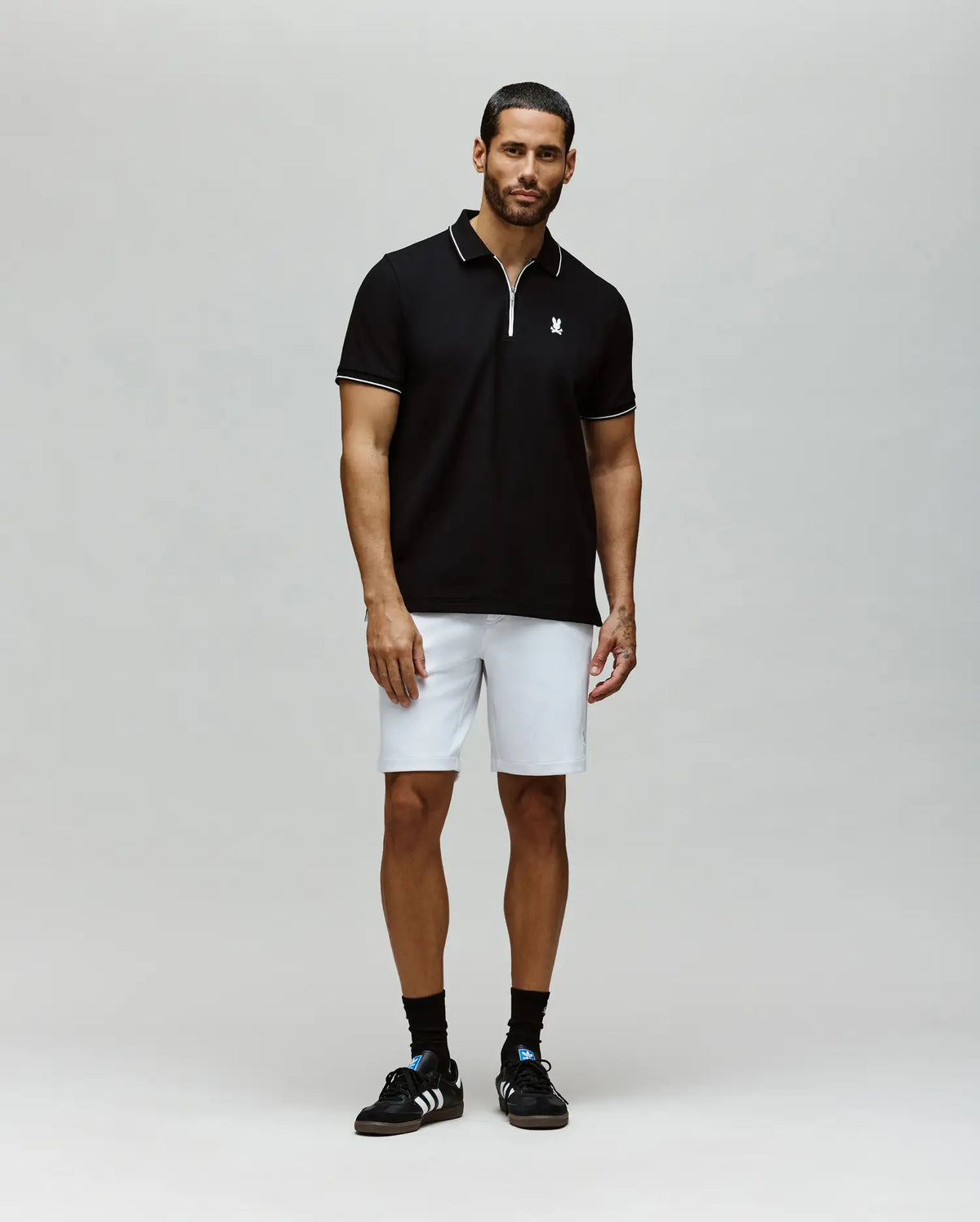 A man with short hair and a groomed beard stands against a plain background wearing the Psycho Bunny MENS DANE ZIP PIQUE POLO - MT0100103, white shorts, black socks, and sneakers. His arms are relaxed at his sides.