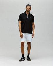 A man with short hair and a groomed beard stands against a plain background wearing the Psycho Bunny MENS DANE ZIP PIQUE POLO - MT0100103, white shorts, black socks, and sneakers. His arms are relaxed at his sides.