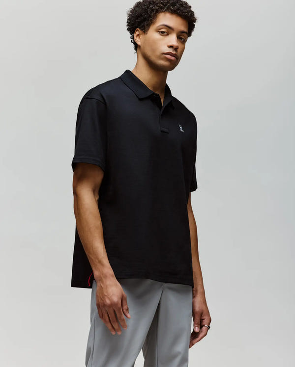 A man with short curly hair wears the Psycho Bunny MENS HENRY OVERSIZED FIT POLO - MT0100104 in black and light gray pants, standing against a plain light background and looking slightly to the side with a neutral expression.