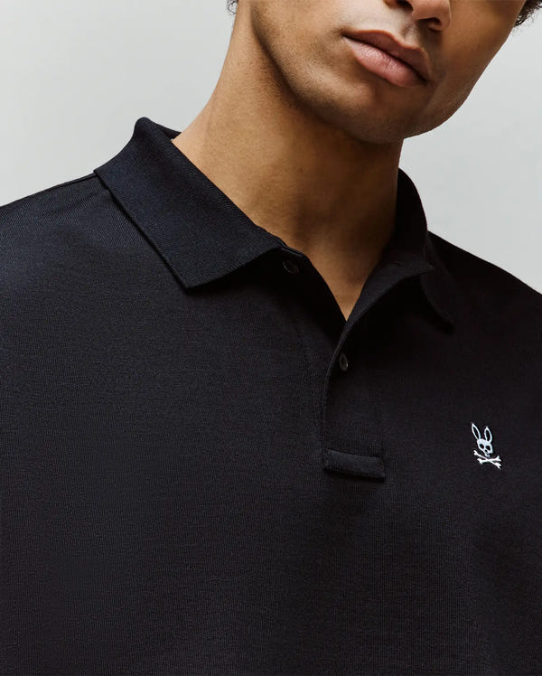 A man wears the Psycho Bunny MENS HENRY OVERSIZED FIT POLO - MT0100104, a heavyweight black Pima cotton shirt with a collared design and a small embroidered Bunny and skull logo on the chest, shown from the shoulders up.