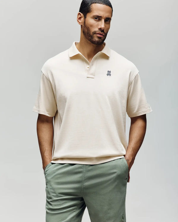 A man with short dark hair and a beard models the Psycho Bunny MENS HENRY OVERSIZED FIT POLO - MT0100104 in cream Pima cotton with a black logo and sage green pants, highlighting its relaxed fit against a plain light background.
