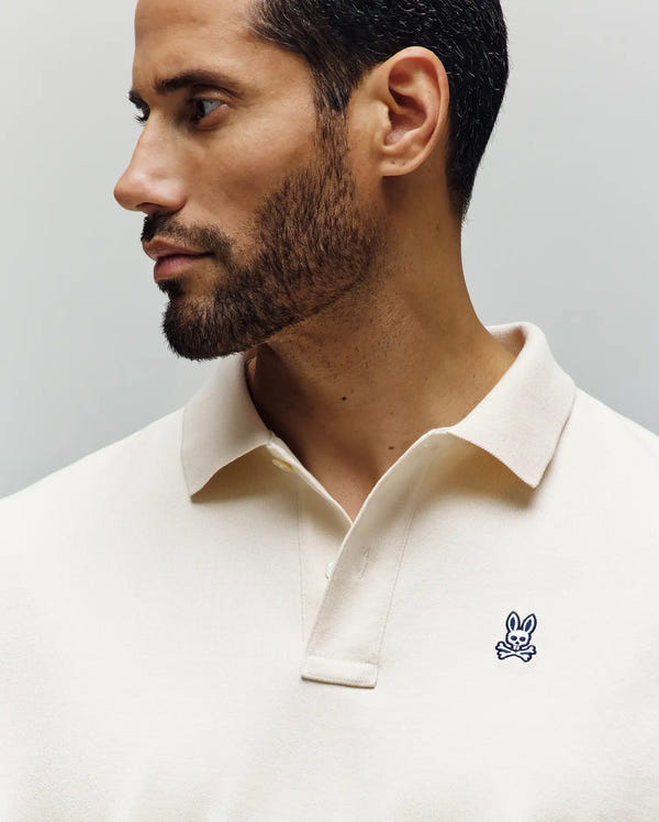 A man with short dark hair and a trimmed beard wears the cream-colored Psycho Bunny MENS HENRY OVERSIZED FIT POLO (MT0100104), made from soft Pima cotton with an embroidered blue rabbit and crossbones logo, looking left against a plain background.